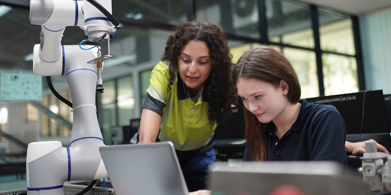 Two women, working in a manufacturing setting, look at a laptop screen.