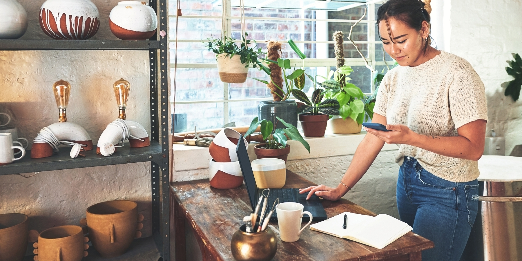 A person uses a phone and laptop at a workbench in a studio filled with ceramics and plants.