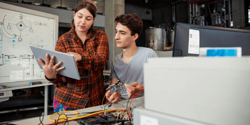 A front view of 2 classmates looking at a digital tablet and helping each other out with coursework in their on-site geothermal engineering plant in Perth, Western Australia.